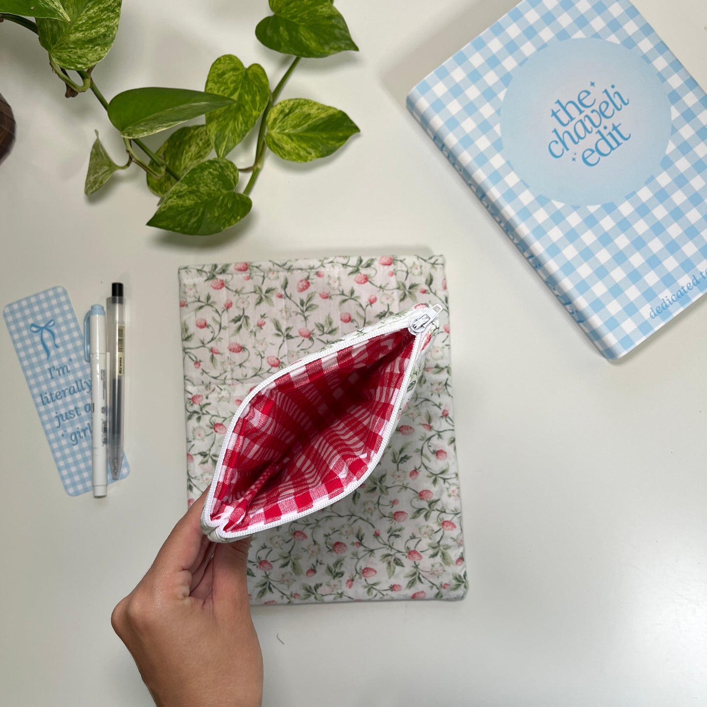 Floral-patterned pouch with a red and white checkered pocket on a white surface, with a plant and book in the background.