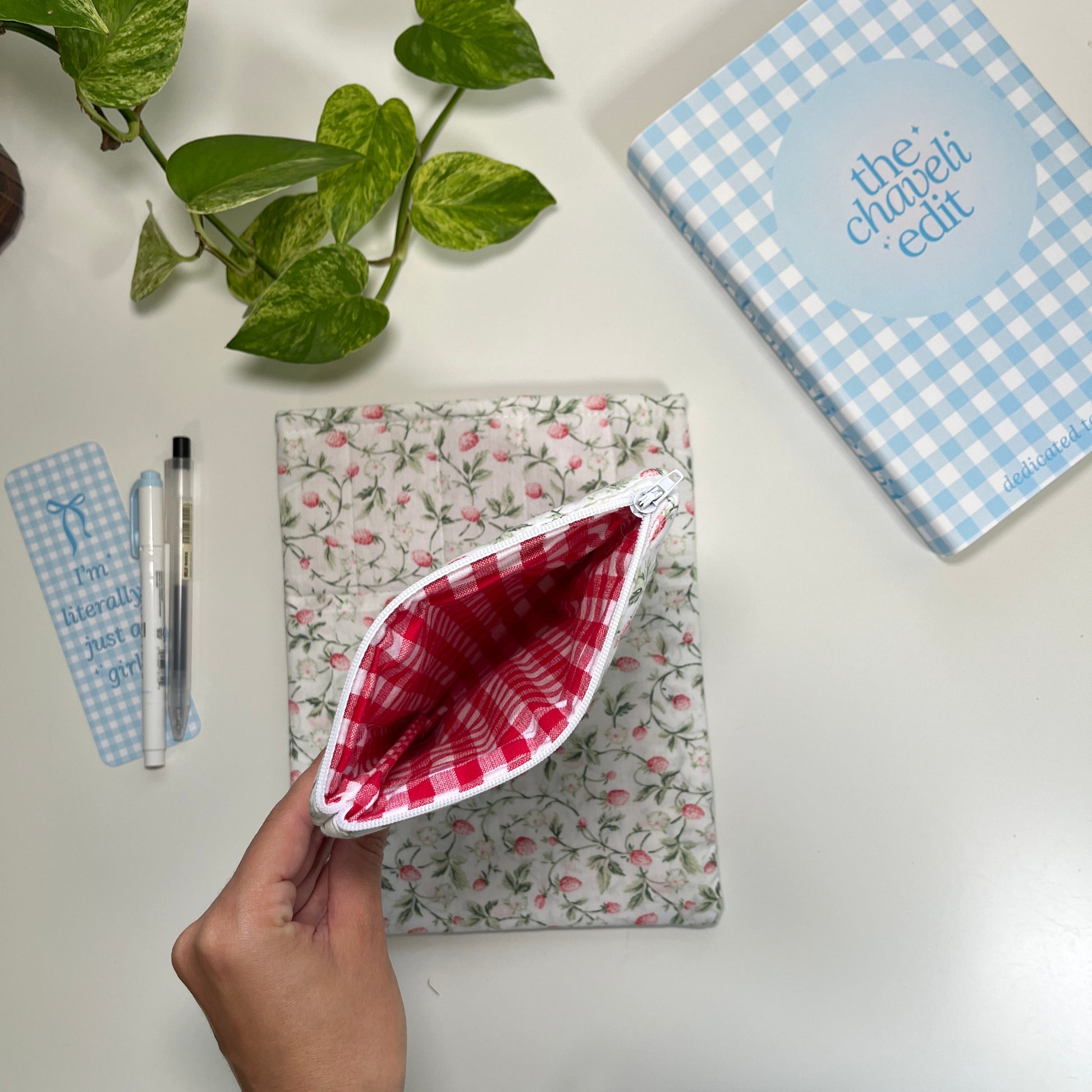Floral-patterned pouch with a red and white checkered pocket on a white surface, with a plant and book in the background.